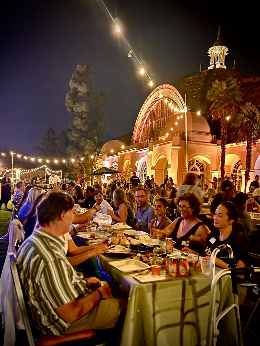Neighbors sharing dinner at one long table beneath string lights and the lit dome of the Botanical Building at the Balboa Park Table
